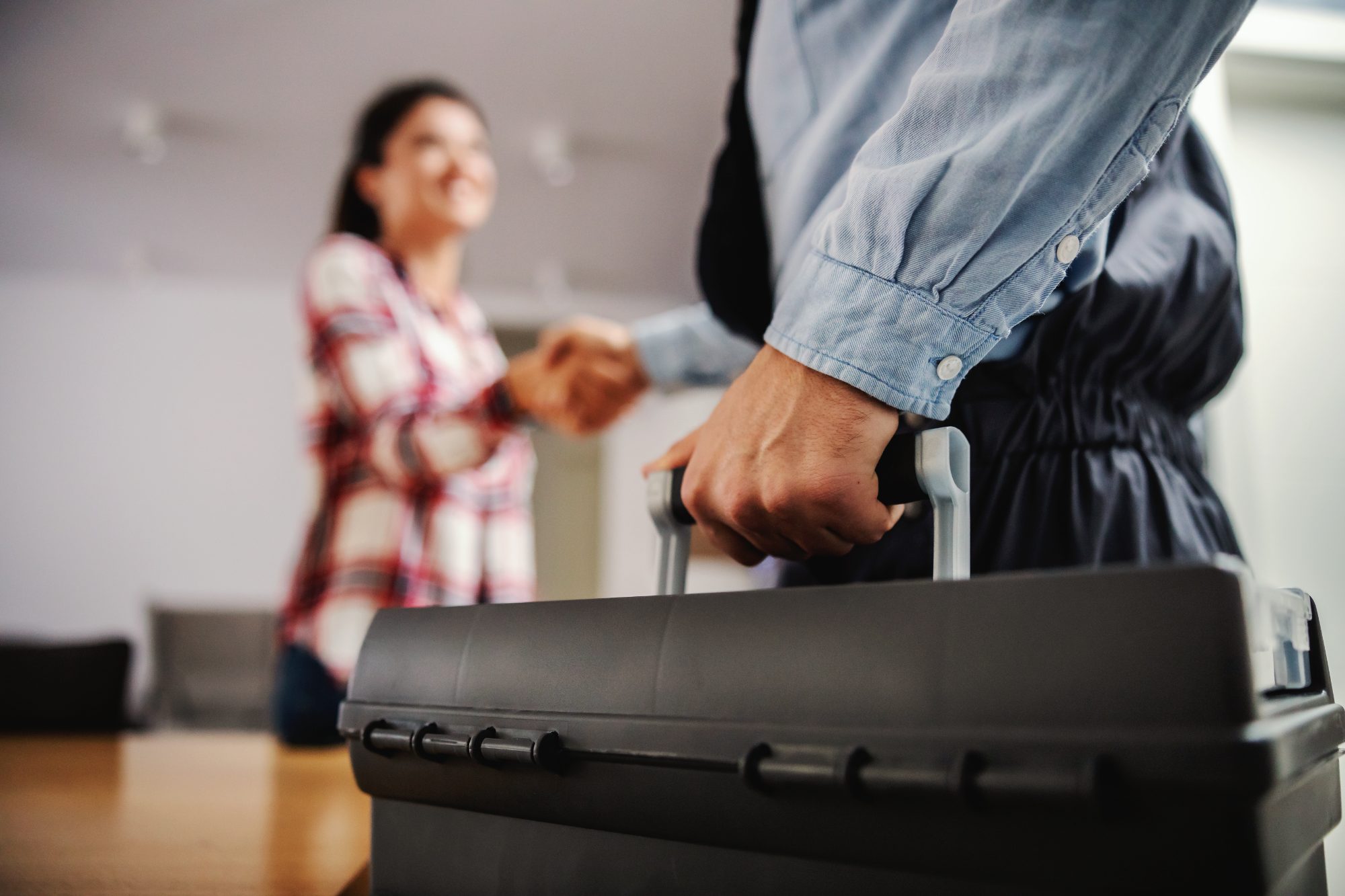 Man in denim shirt pulling handle of black rolling suitcase while woman in plaid shirt stands in background